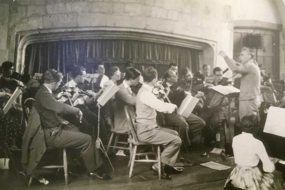An old photo of a man conducting an orchestra, full of string musicians, with a female accompanist next to the composer.